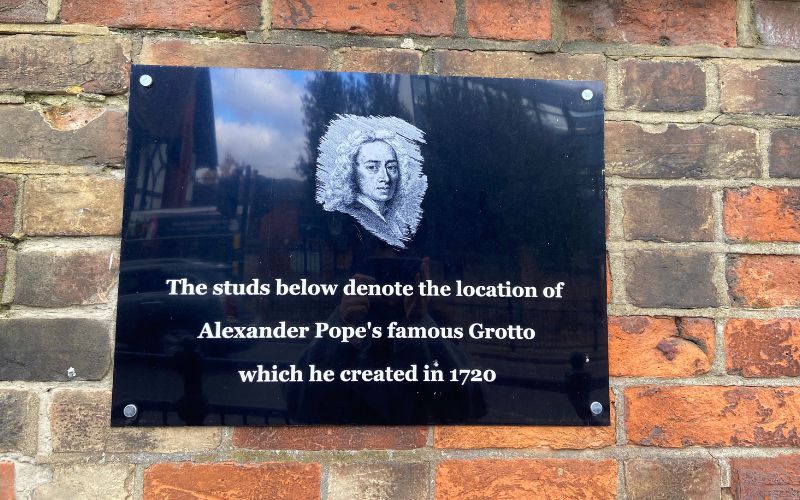 Plaque outside of Alexander Pope's Grotto. On a brick wall. Is black with white writing saying "The studs below denote the location of Alexander Pope's famous Grotto which he created in 1720." And above the writing is an etching of Alexander Pope.