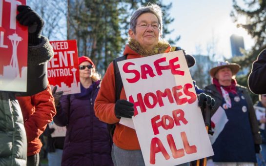 Elderly woman at a protest with a sign in red that says: 'Safe homes for all'
