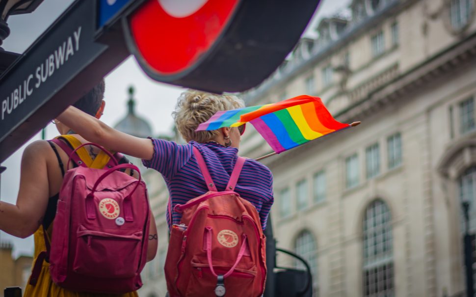 A couple stands next to the London underground, holding their arms around one another and holding a pride flag.