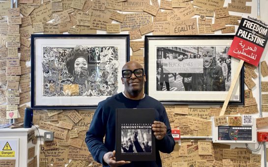 Wayne Campbell is holding his book 'A celebration of demonstration'. He is standing in front of a wall with two of his photographs. The wall is covered with pieces of cardboard featuring quotes and comments