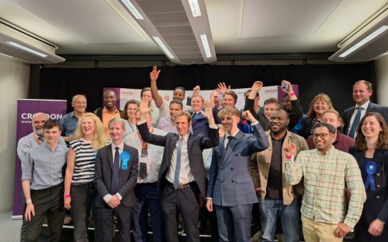 Chris Philp poses with his arms raised in front of a group of Conservatives