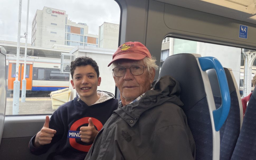 Picture shows young boy and his grandad sat aboard a train. The young boy has his thumbs up to the camera. They are smiling.