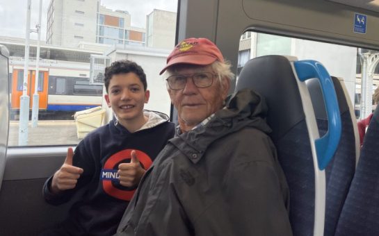 Picture shows young boy and his grandad sat aboard a train. The young boy has his thumbs up to the camera. They are smiling.