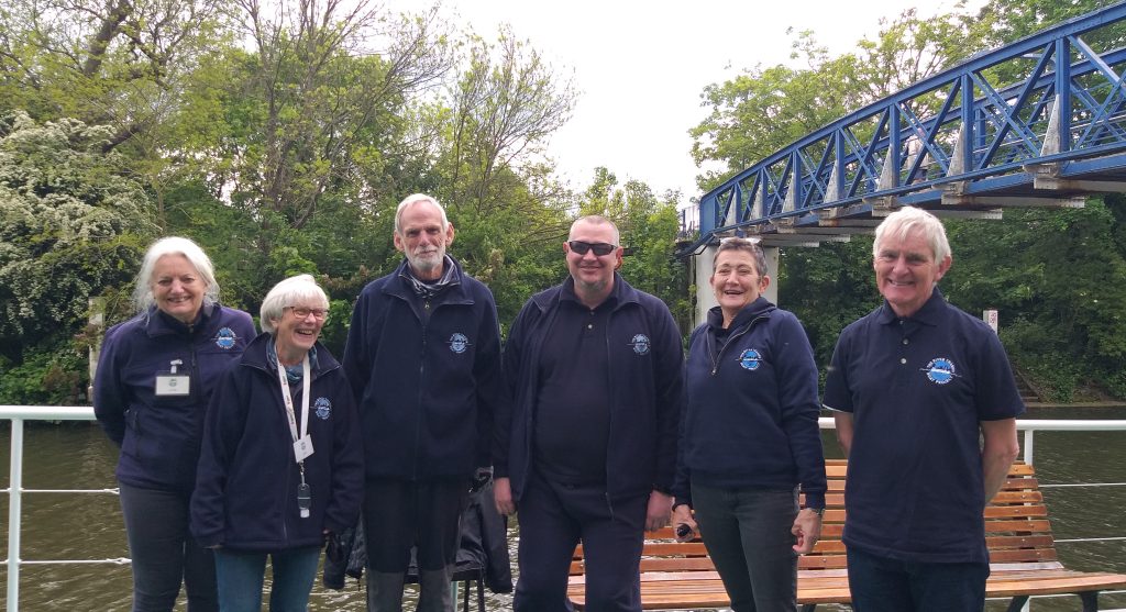 Six people smiling at the camera on board a boat. They are each wearing navy blue tops.