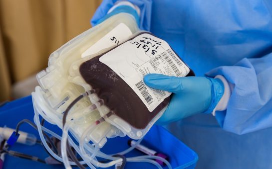 A person wearing medical overalls and gloves holds a package of donated blood