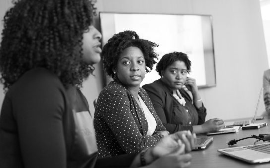 grayscale photography of two women on conference table looking at talking woman
