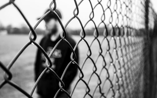 A black and white image showing a person standing behind a fence. The individuals silhouette is visible against the patterned lines of the barrier, conveying a sense of separation and confinement.