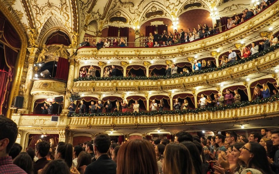 A wide angle shot of the inside of an opera theatre, looking from the stalls up towards the balconies. There is a full audience. They are giving a standing ovation.