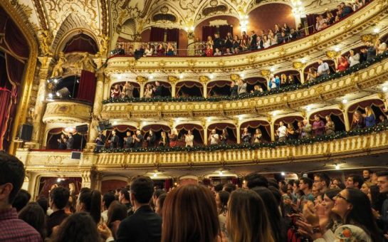 A wide angle shot of the inside of an opera theatre, looking from the stalls up towards the balconies. There is a full audience. They are giving a standing ovation.