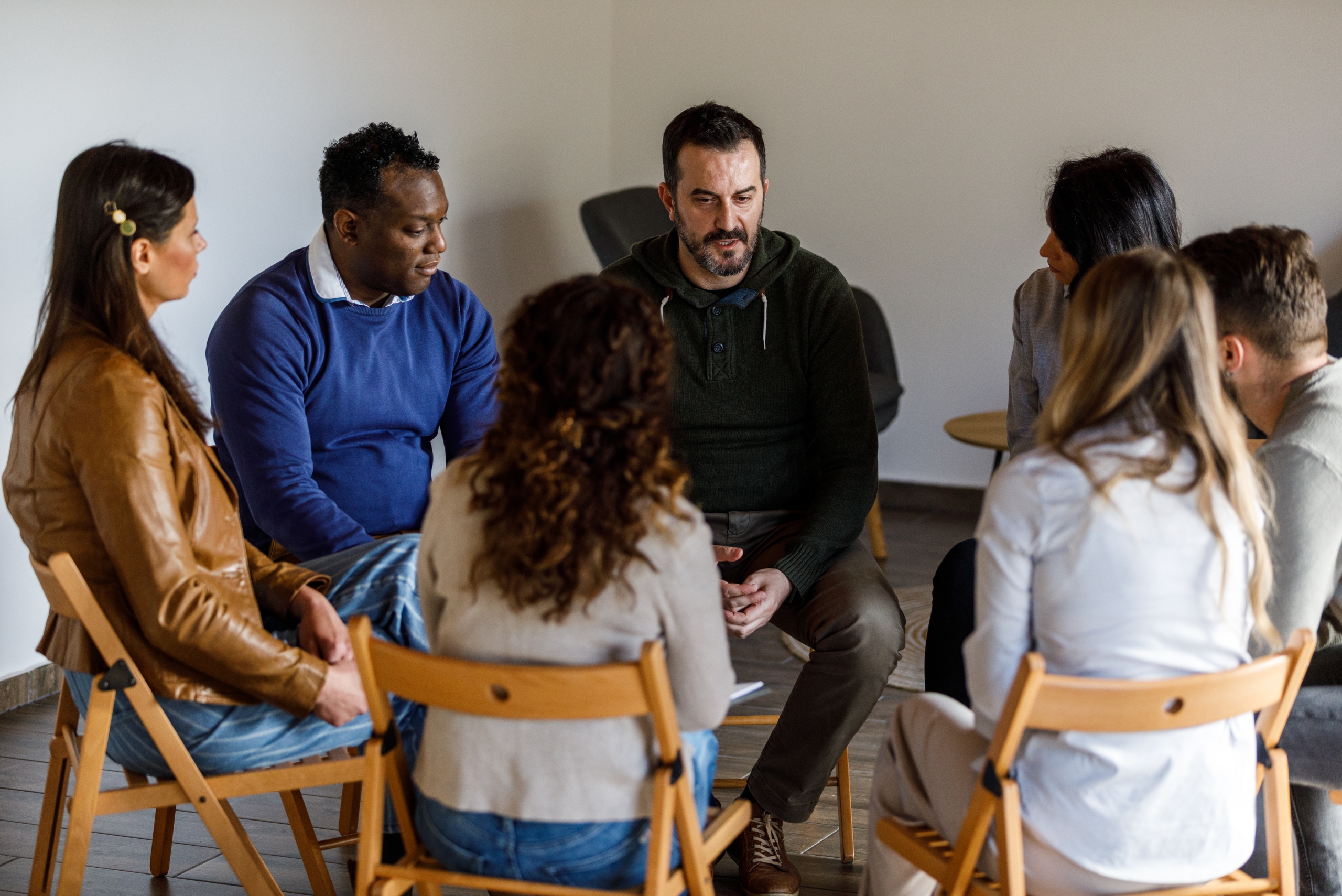 A group of seven adults sit in a circle on wooden fold out chairs in conversation