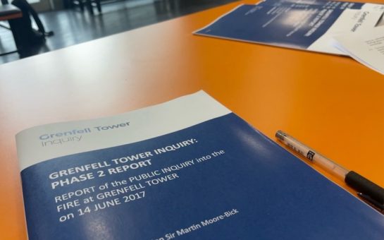 A blue and white booklet is on its title page and concerns Stage 2 of the Grenfell Tower Inquiry. It sits on a large orange table.