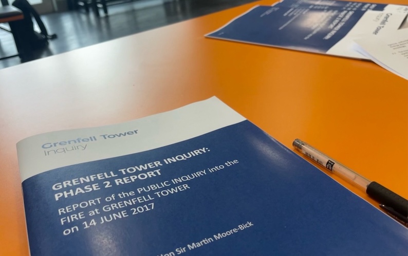 A blue and white booklet is on its title page and concerns Stage 2 of the Grenfell Tower Inquiry. It sits on a large orange table.