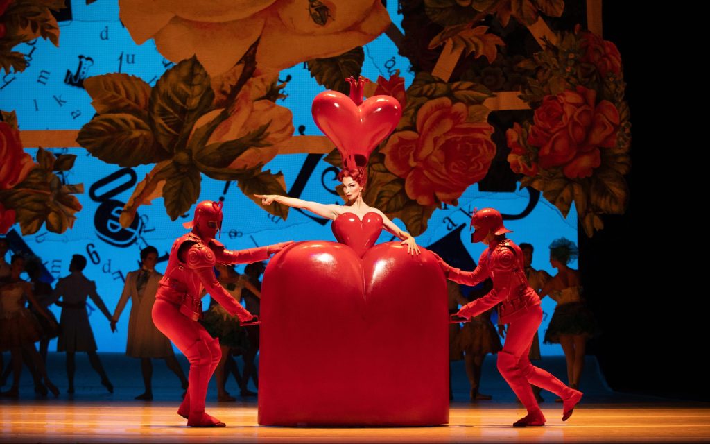 The Queen of Hearts pointing to the left in her wooden dress-shaped cart being wheeled across the stage by two servants