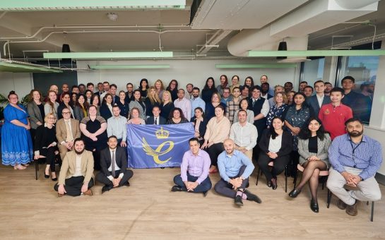 The staff at FACT are arranged around a flag containing The King's Award logo in a large room. Some are sat on the floor some are stood.