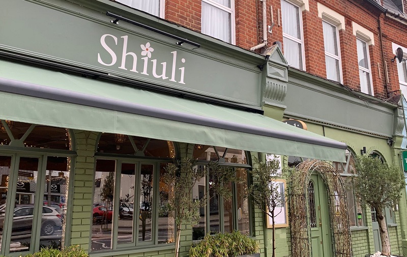 Image of the outside of Shiuli. the green outside with large windows and olive trees outside the entrance