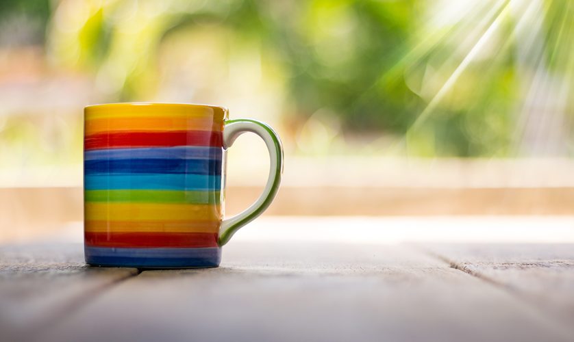 A mug of tea with the rainbow flag painted on it sits on a wooden table.