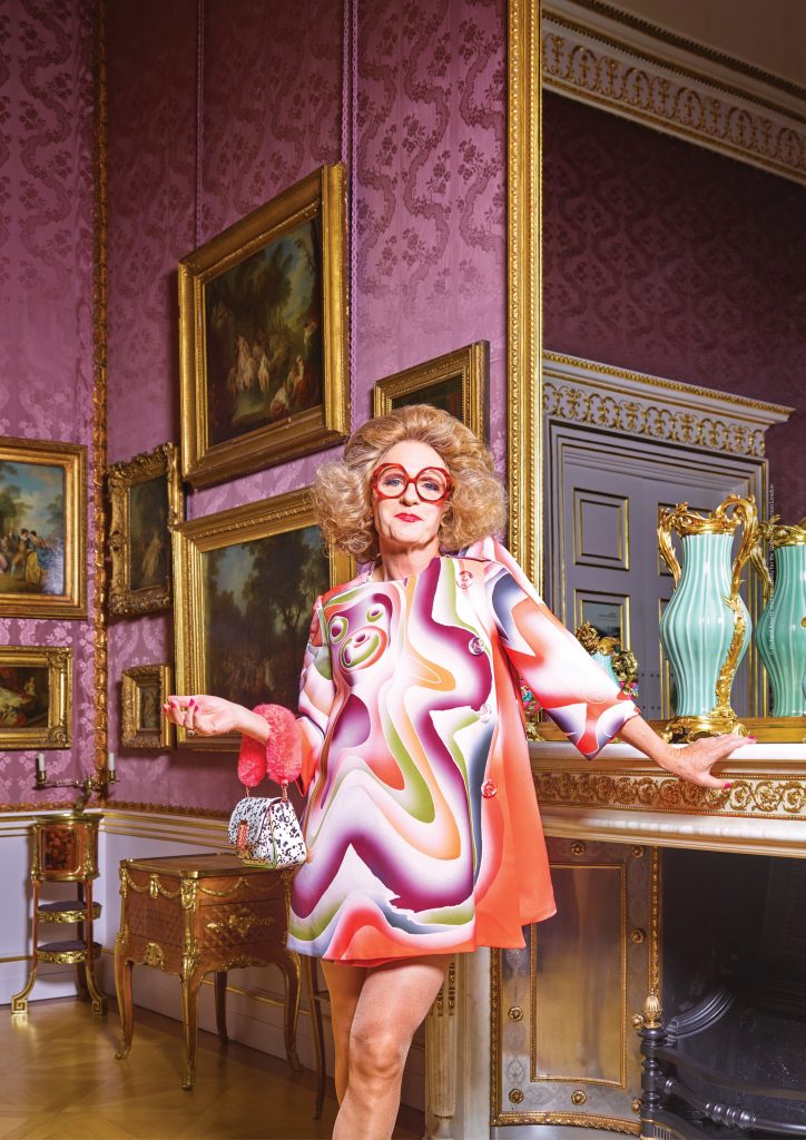 Image of the artist Grayson Perry in a 70s style dress in a Wallace Collection room by a mirror