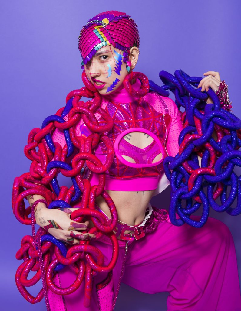 Person in pink outfit with red and blue material chain worn as  a scarf