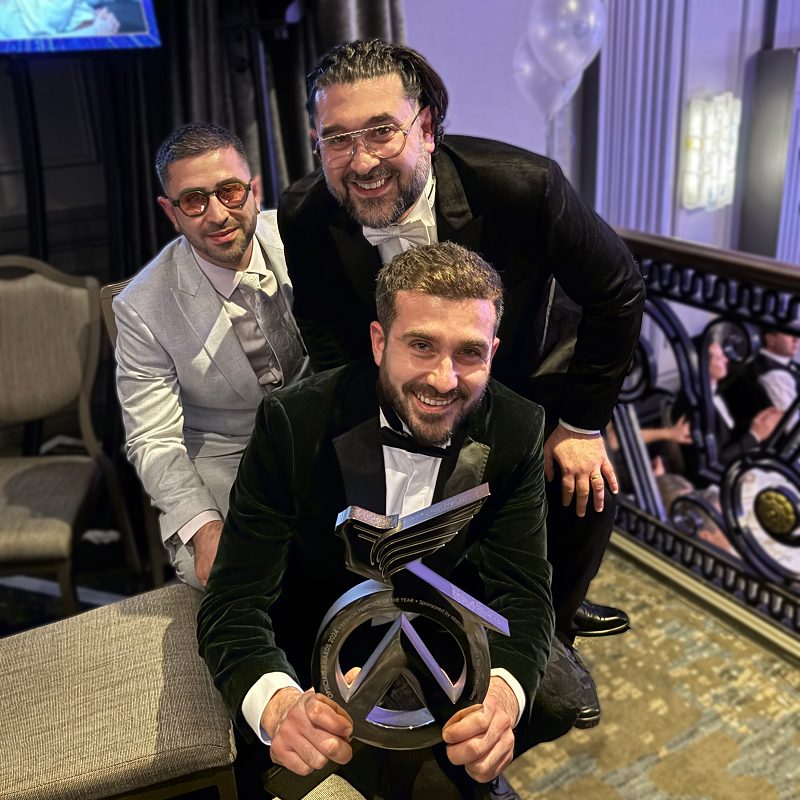 The image shows the three brothers dressed in formal attire, smiling and posing together at an event. The man in the front, wearing a black tuxedo, is holding an Opticians Award. The two men behind him are also wearing formal suits—one in black and the other in light grey. The setting appears to be an elegant venue with decorative railings and a festive atmosphere.