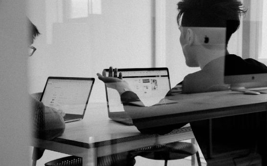 Two men with laptops sat behind a glass partition, having a meeting and discussion.