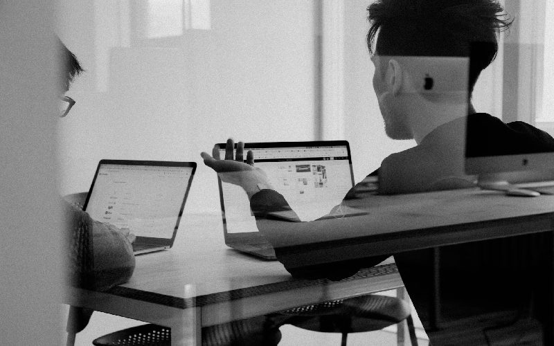 Two men with laptops sat behind a glass partition, having a meeting and discussion.