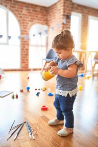 A female toddler plays with a toy. There are many other toys and paintbrushes scattered around her feet and some blue bunting on the wall behind her.