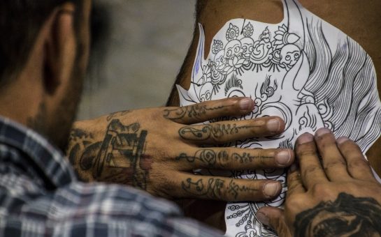 A person with tattooed hands holds a tattoo stencil to their leg.